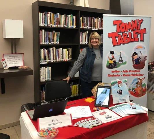 Julie Prantera smiling at her author table in a library, with the Tommy Tablet banner, copies of both books, coloring sheets, and a tablet on a red tablecloth.