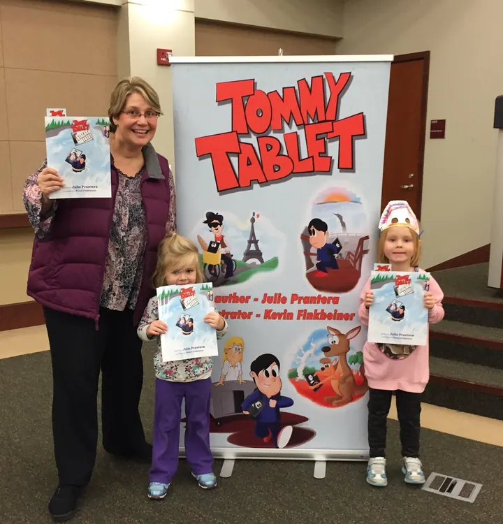 Julie Prantera smiling beside the Tommy Tablet banner with two young readers, all holding Tommy Tablet at Niagara Falls.